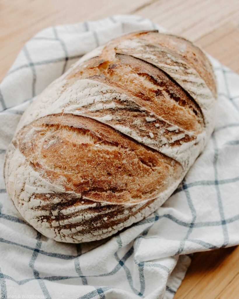 Rustic, golden-brown loaf of sourdough bread on a white and blue checkered cloth. The loaf has a crusty texture, conveying warmth and freshness.