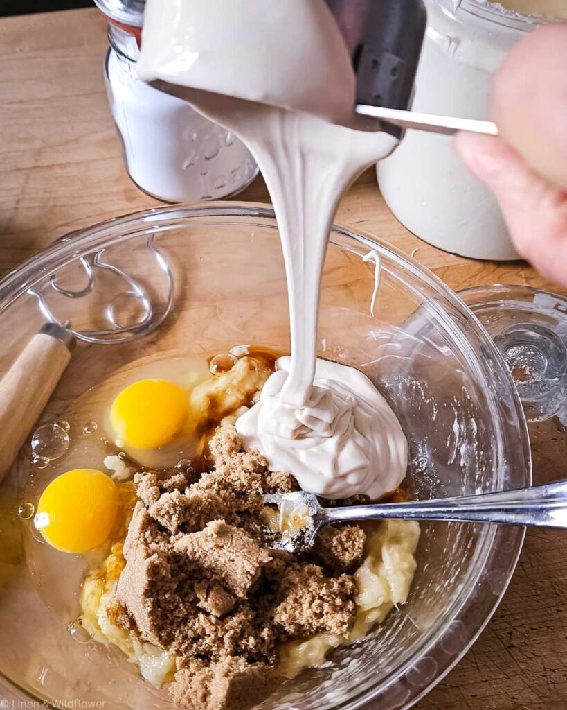 Sourdough discard is being poured into a mixing bowl with eggs, brown sugar, and mashed bananas on a wooden countertop, evoking a cozy baking scene.