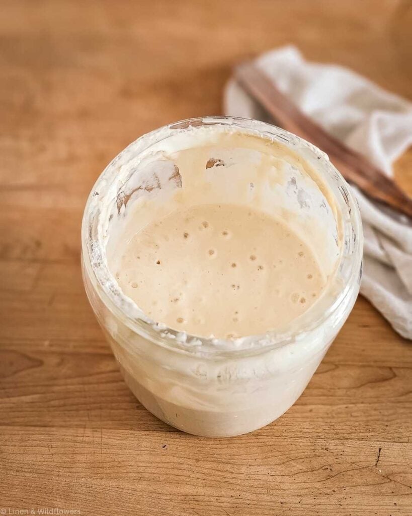 A glass jar filled with creamy sourdough starter, slightly bubbly, sits on a wooden surface. A beige cloth is partially visible in the background.