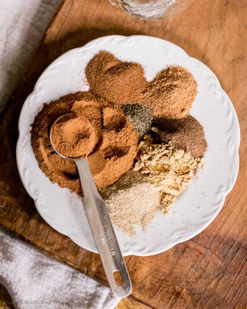 A white plate on a wooden table holds piles of various powdered spices, including cinnamon and ginger, with a metal teaspoon resting on top. Cozy and warm tones.