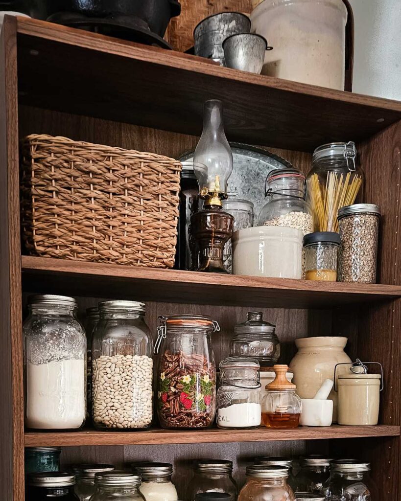 Wooden pantry shelves hold jars of pasta, grains, beans, herbs, and kitchenware. A cozy and rustic vibe with a woven basket and vintage lamp.