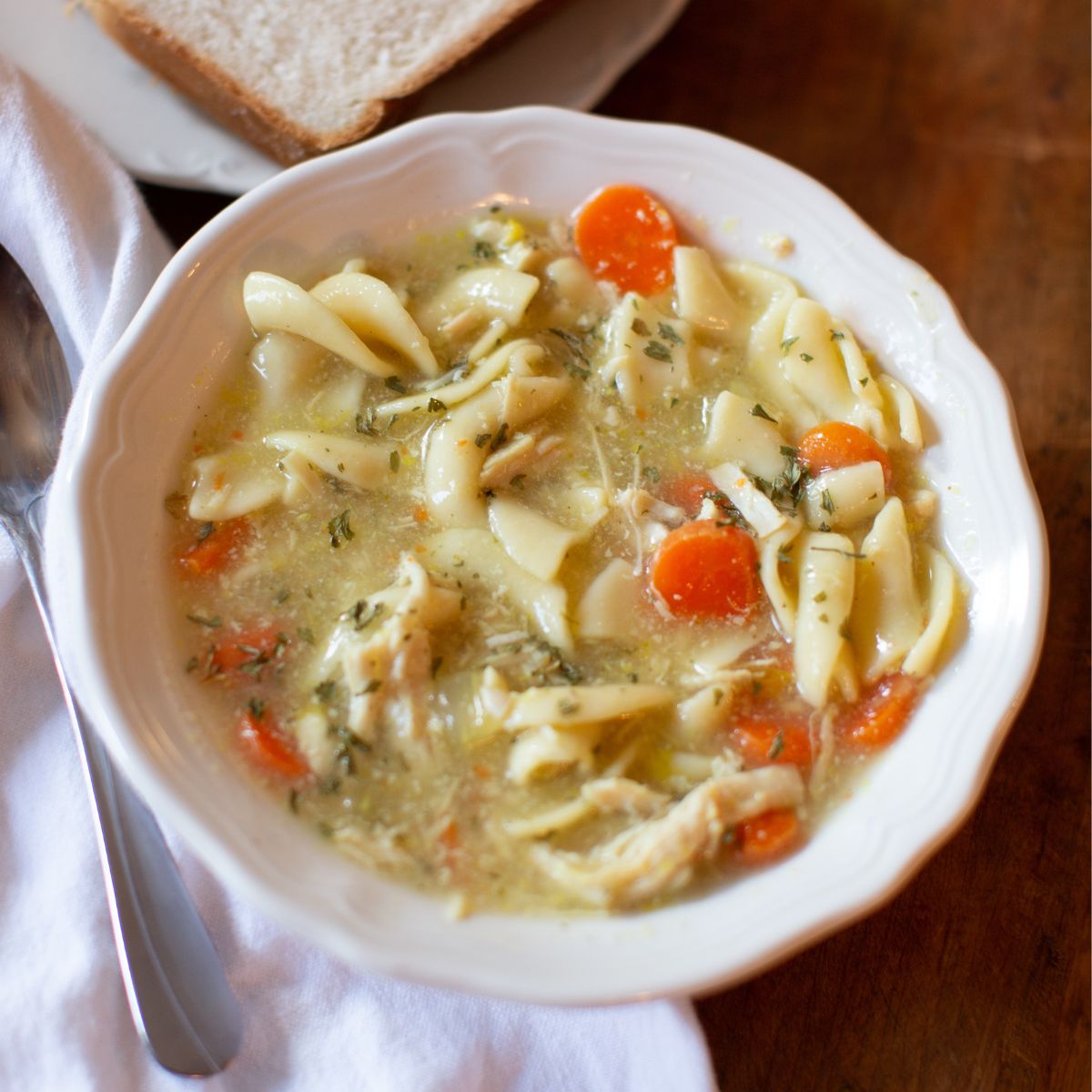 A bowl of chicken noodle soup with carrots and herbs sits on a wooden table beside slices of white bread. The scene conveys comfort and warmth.