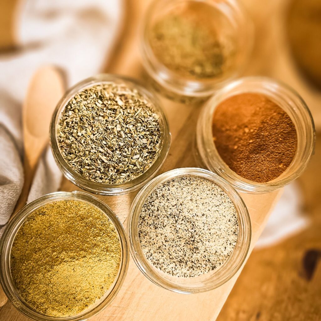 Five small glass jars filled with various spices are arranged on a wooden surface. Each jar holds spices of different colors and textures, creating a warm, inviting atmosphere.
