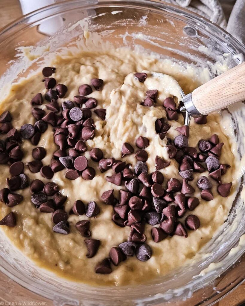 A glass bowl filled with creamy banana bread batter topped with a generous amount of chocolate chips. A wooden spoon is partially visible, suggesting baking in progress.