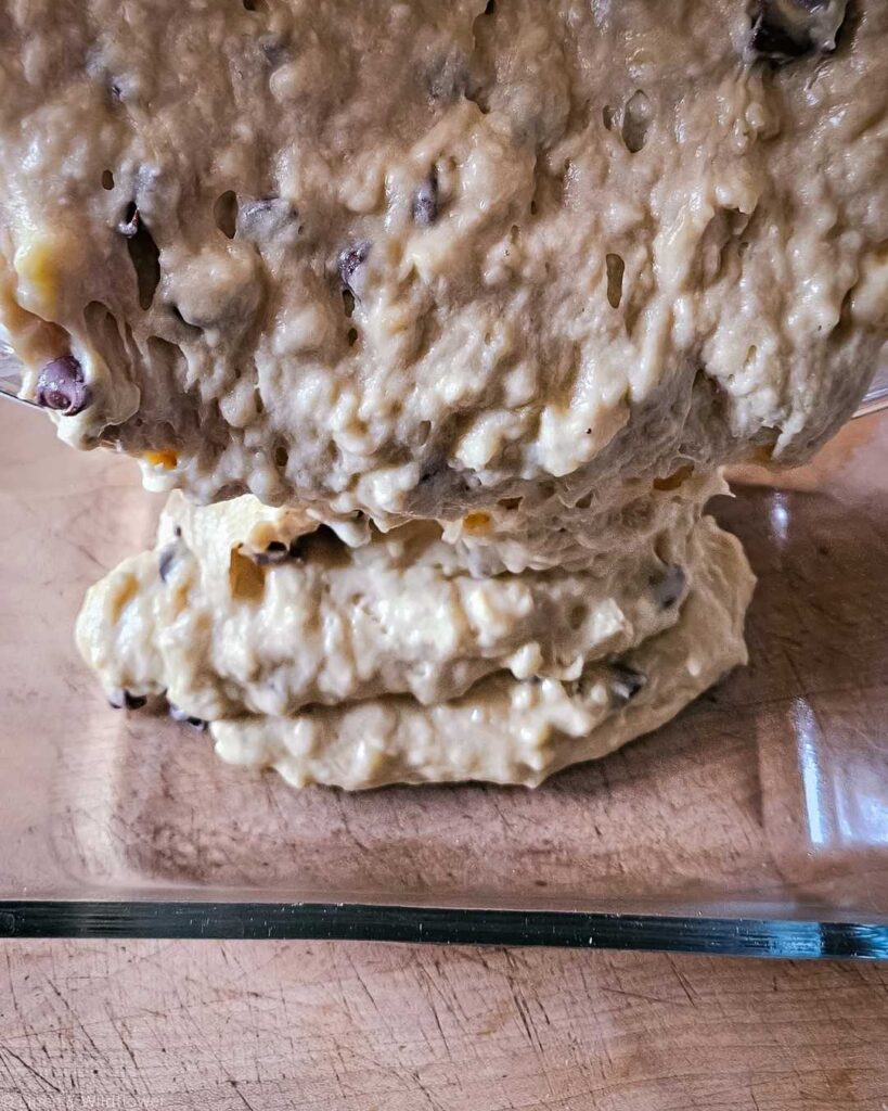 A glass bowl filled with creamy banana bread batter topped with a generous amount of chocolate chips. A wooden spoon is partially visible, suggesting baking in progress.