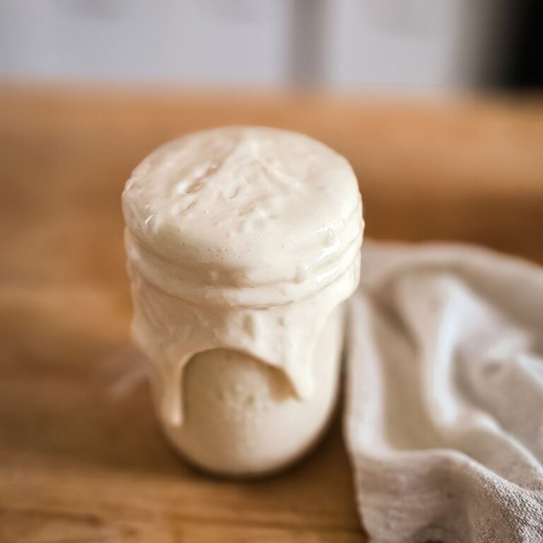 A mason jar overflowing with creamy, pale batter sits on a wooden table. A cloth rests beside it, suggesting baking or cooking preparation.