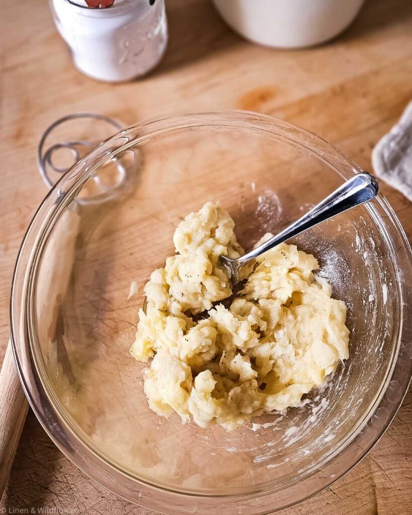 A glass bowl on a wooden surface contains creamy, mixed banana batter, with a spoon. Nearby are flour and sugar containers, creating a cozy, baking atmosphere.