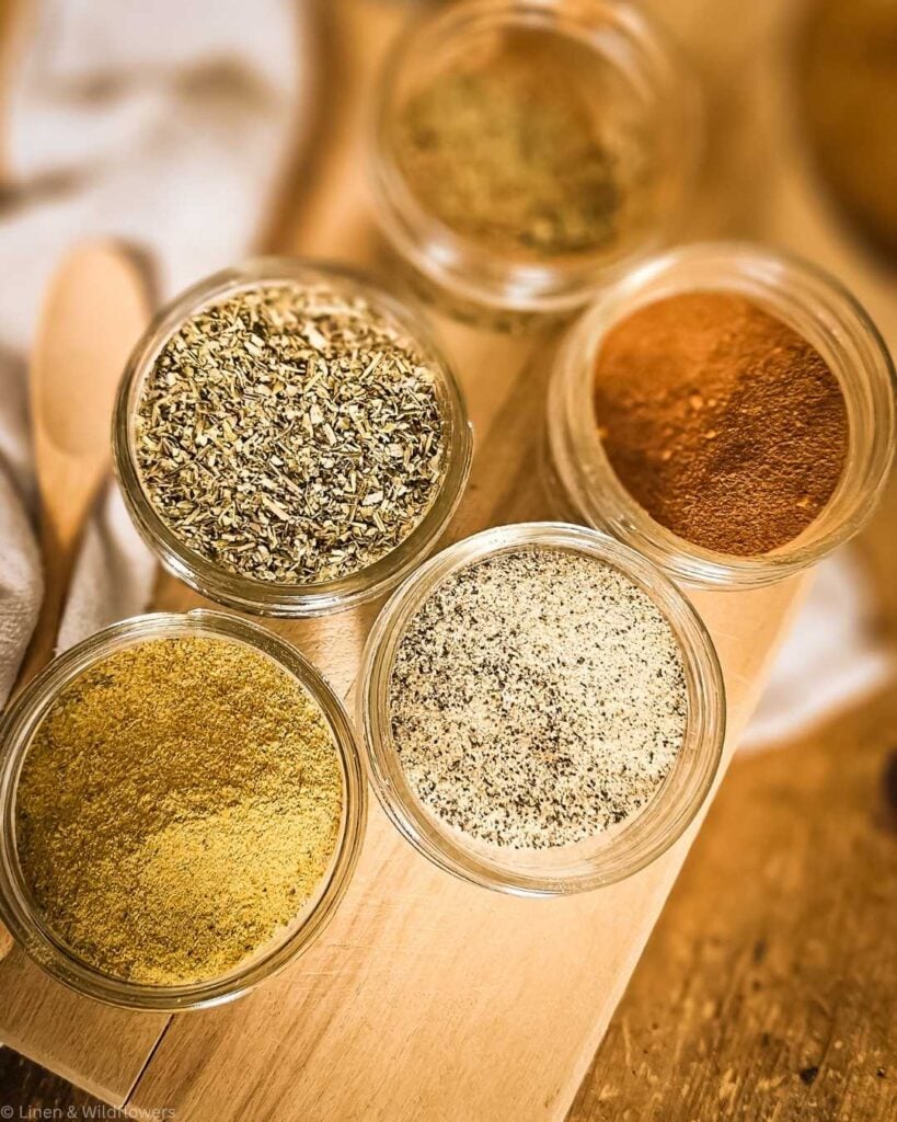 Overhead view of five small glass jars filled with various spices on a wooden board. Warm tones and textures create a cozy, aromatic atmosphere.