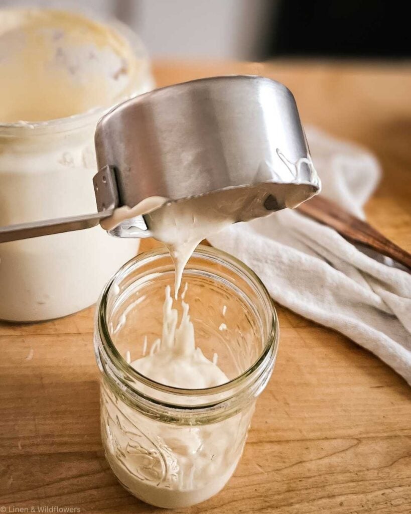Sourdough starter liquid is being poured from a silver measuring cup into a glass jar on a wooden table, with a cloth and container in the background.