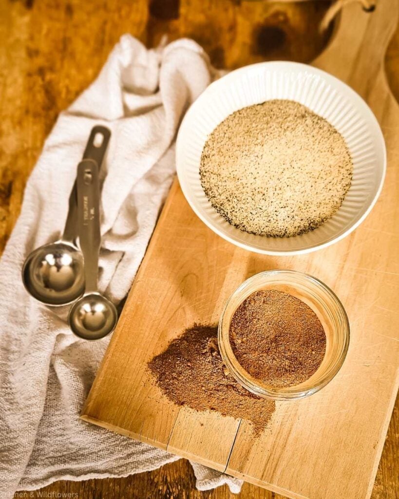 A wooden cutting board holds a white bowl of sesame seeds and a jar of ground spices, with spilled spices nearby. Metal measuring spoons and a white towel add a rustic feel.