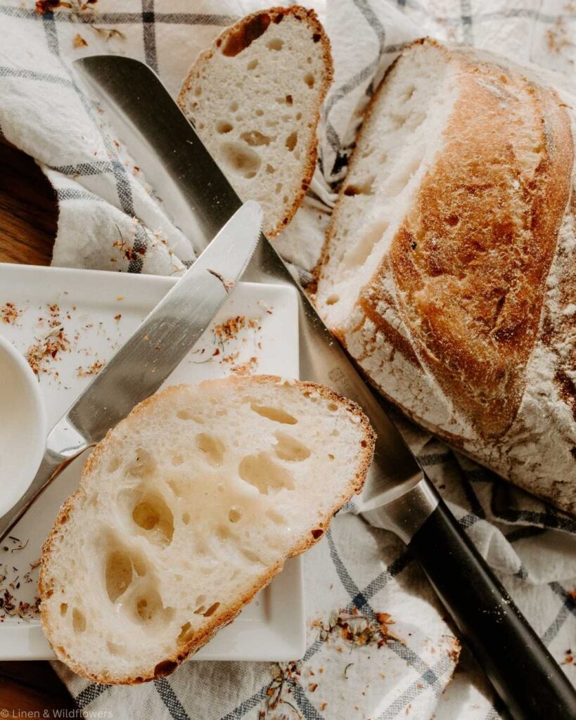 A sliced loaf of rustic bread on a checkered cloth. A knife rests next to the bread, conveying a cozy, artisanal kitchen vibe.