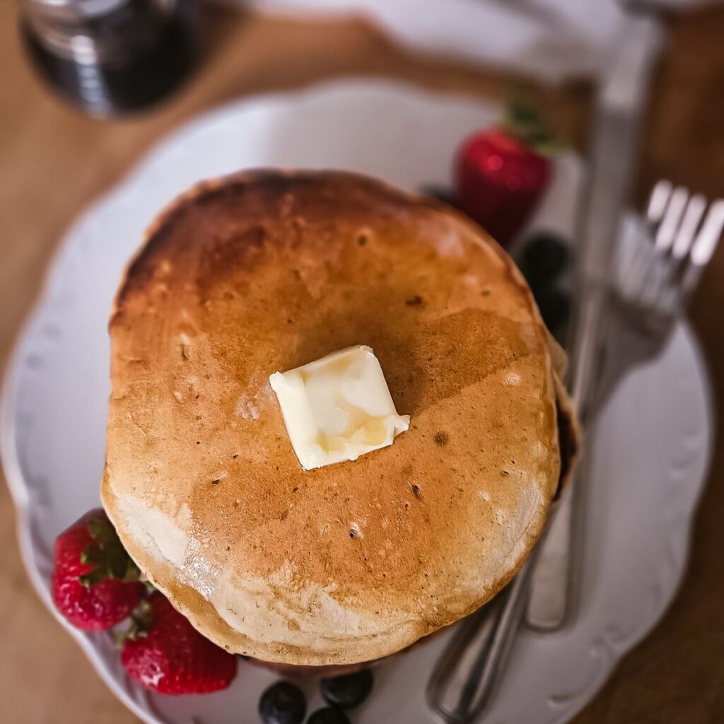 A stack of golden sourdough pancakes topped with a melting butter pat, served with fresh strawberries and blueberries. A fork and a knife are placed beside it.