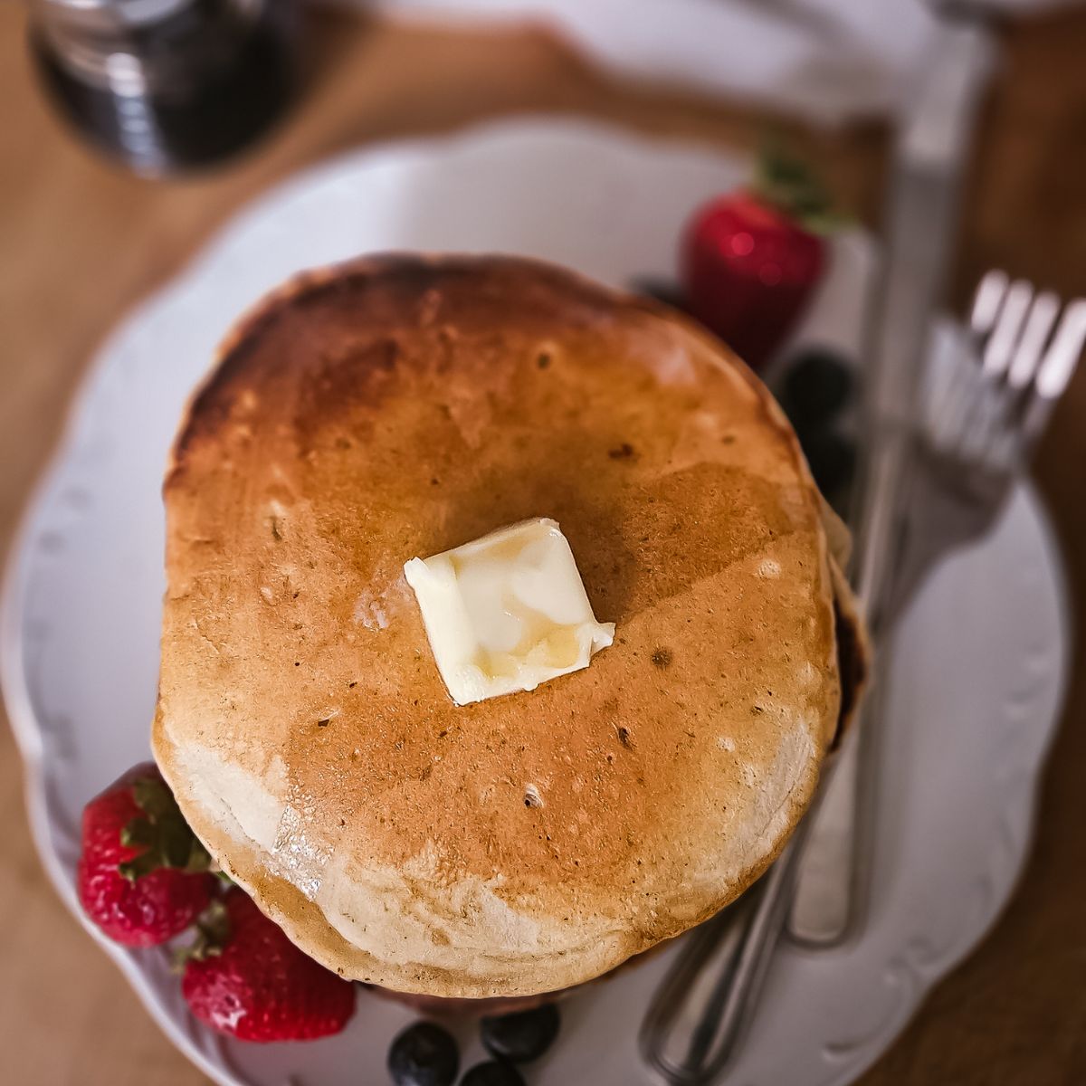 A stack of golden sourdough pancakes topped with a melting butter pat, served with fresh strawberries and blueberries. A fork and a knife are placed beside it.