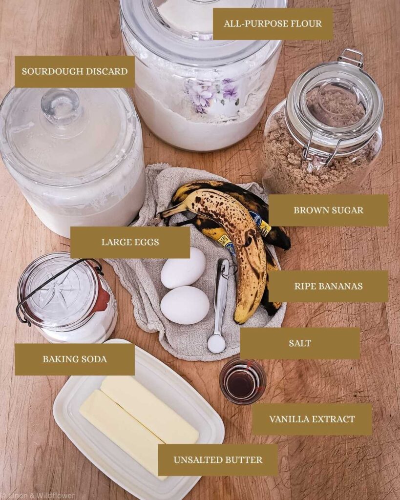 Ingredients for baking are arranged on a wooden table: all-purpose flour, sourdough discard, brown sugar, ripe bananas, eggs, butter, baking soda, salt, and vanilla.