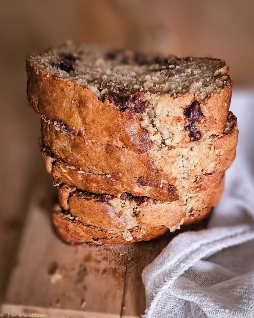 Three thick slices of banana bread with dark chocolate chunks stacked on a wooden board. The warm tones convey a cozy, homemade feel.