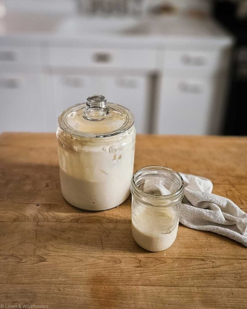 Glass jar filled with creamy sourdough starter next to a smaller jar on a wooden countertop. A white cloth lies nearby, creating a cozy kitchen vibe.