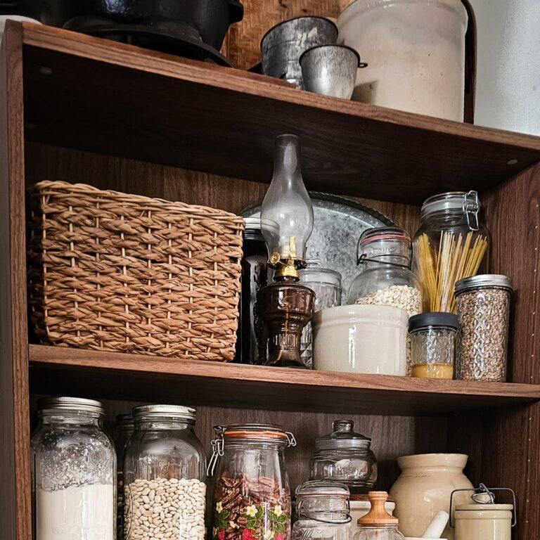 Wooden pantry shelves hold jars of pasta, grains, beans, herbs, and kitchenware. A cozy and rustic vibe with a woven basket and vintage lamp.