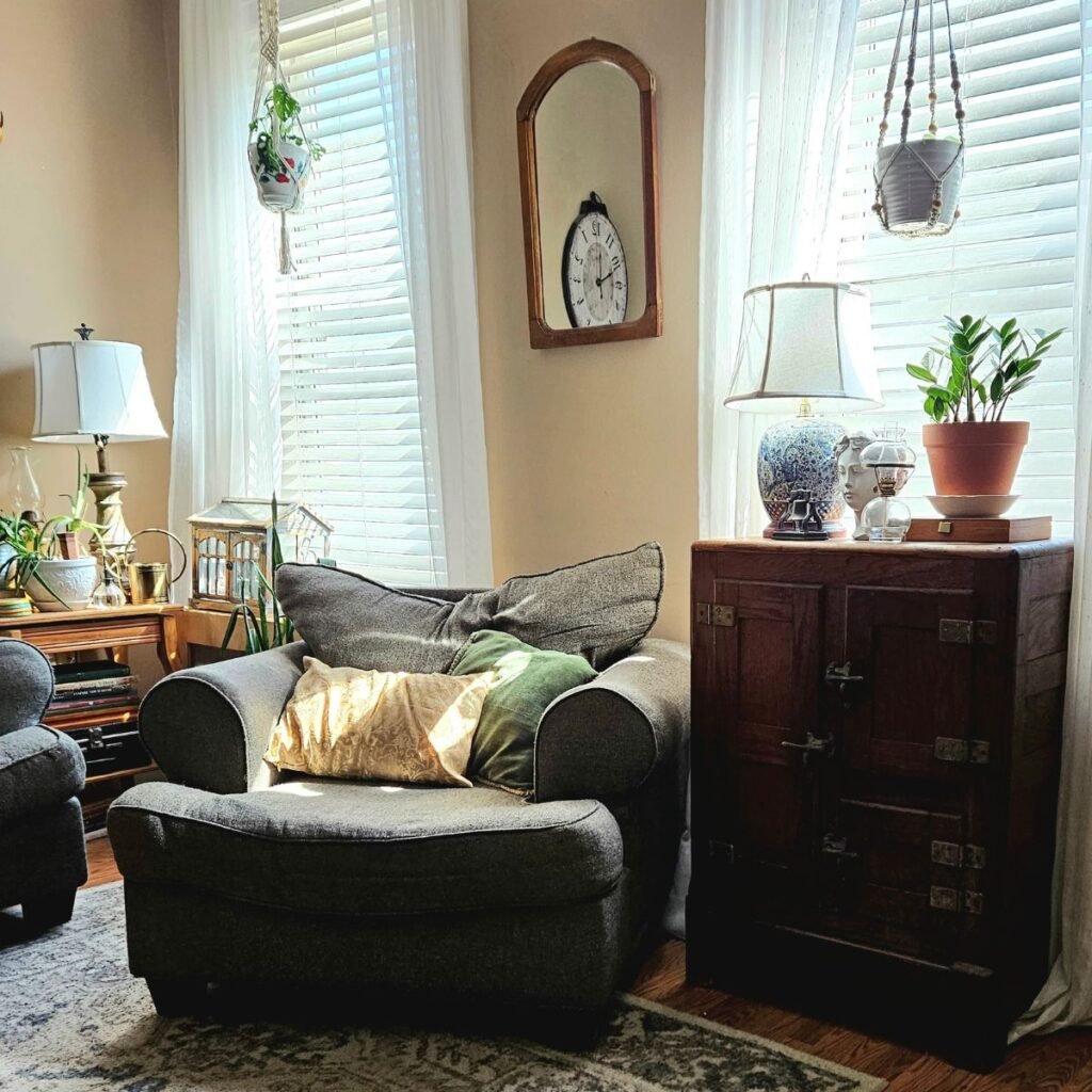 Cozy living room with a gray armchair, warm sunlight, and cushions. Wood cabinet with a lamp and plant. Hanging plants by two tall windows. Relaxed ambiance.