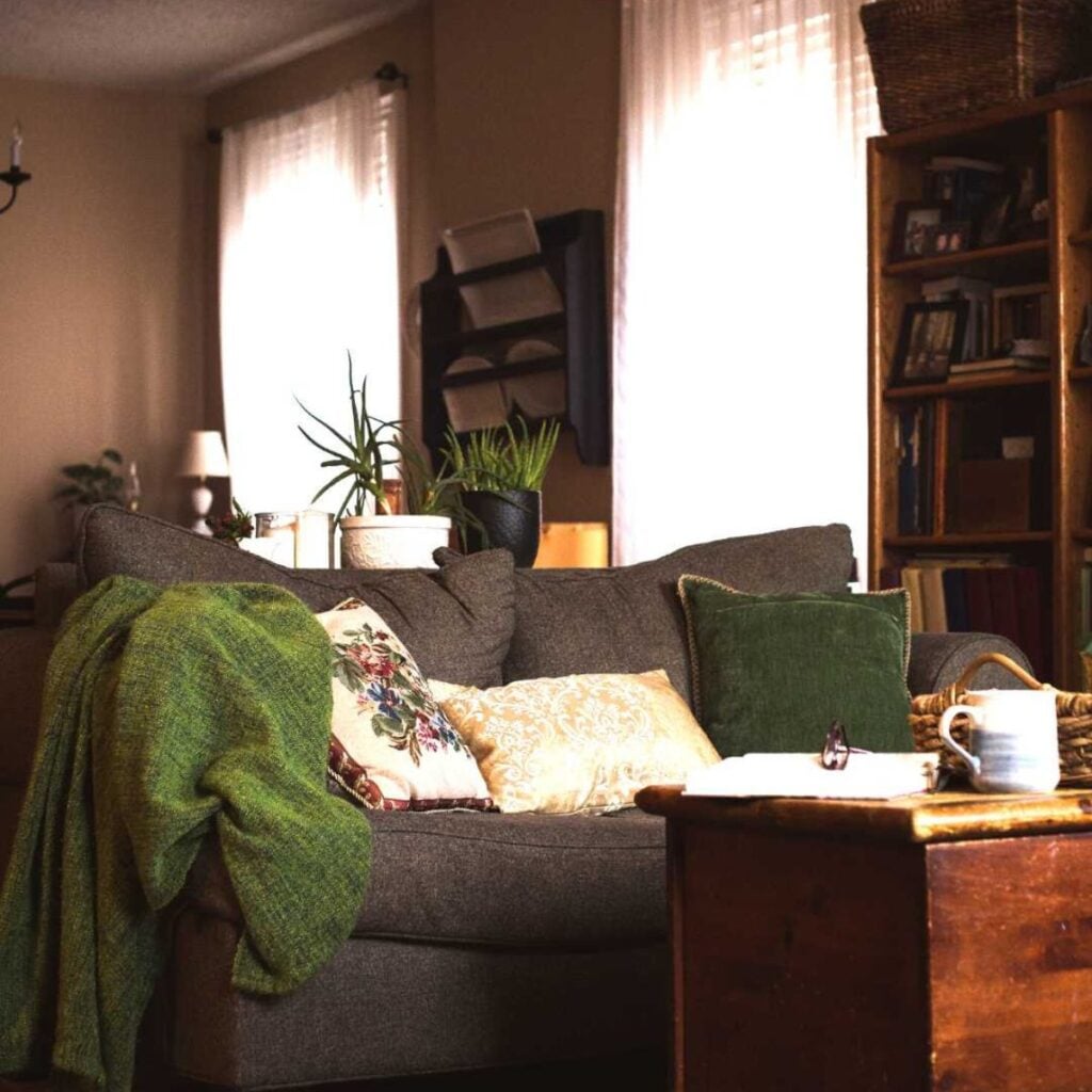 Cozy living room with a gray sofa adorned with green and floral pillows, a green throw blanket, and potted plants. A wooden coffee table holds a mug and papers, while soft light filters through white curtains, creating a warm and inviting atmosphere.