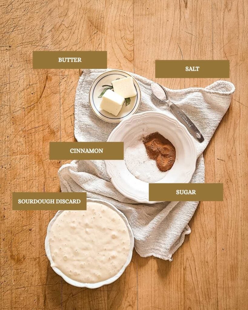 Wooden table with labeled baking ingredients: sourdough discard in a bowl, butter blocks, sugar with cinnamon mix, and salt on a metal spoon. Rustic theme.