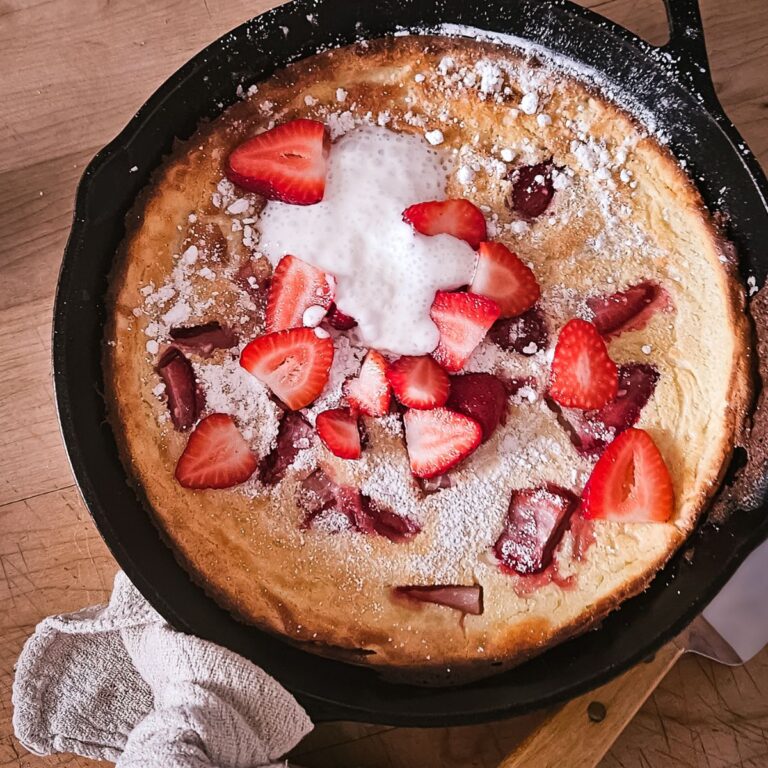 Golden Dutch baby pancake in a skillet, topped with fresh strawberry slices, powdered sugar, and a dollop of cream, on a wooden table.