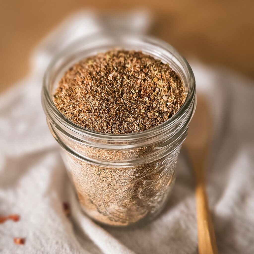 A glass mason jar filled with a colorful homemade taco seasoning blend on a white cloth, with a wooden spoon nearby. The warm tones convey a cozy, inviting atmosphere.