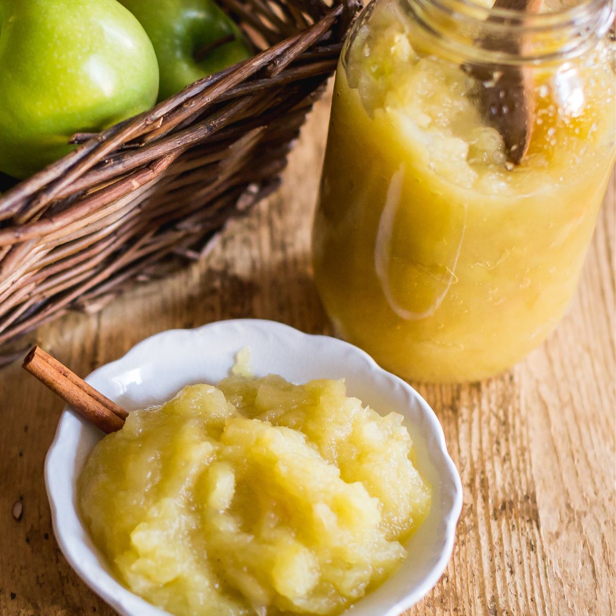 Basket of green apples next to bowls and a jar of applesauce with cinnamon sticks on a wooden table. The scene is warm and rustic.