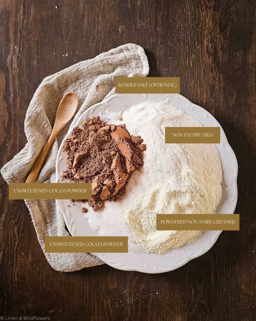 White plate holds piles of unsweetened cocoa, non-fat dry milk, and powdered non-dairy creamer on a dark wooden table. A towel and spoon lie nearby.