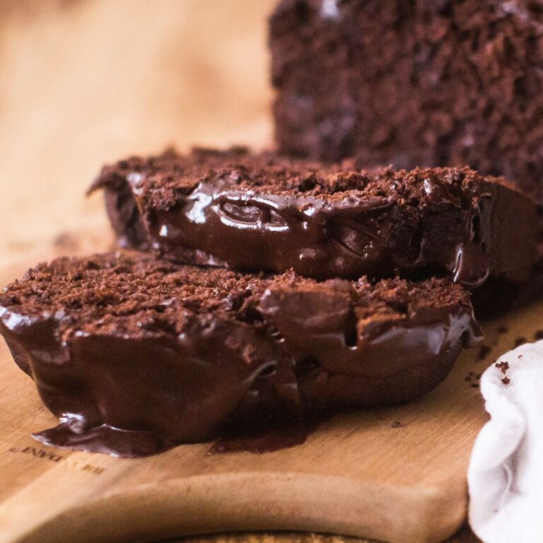 Close-up of a sliced, moist chocolate cake on a wooden cutting board. The cake's rich texture and glossy glaze evoke indulgence. A floral plate and white cloth are partially visible, adding a cozy touch.