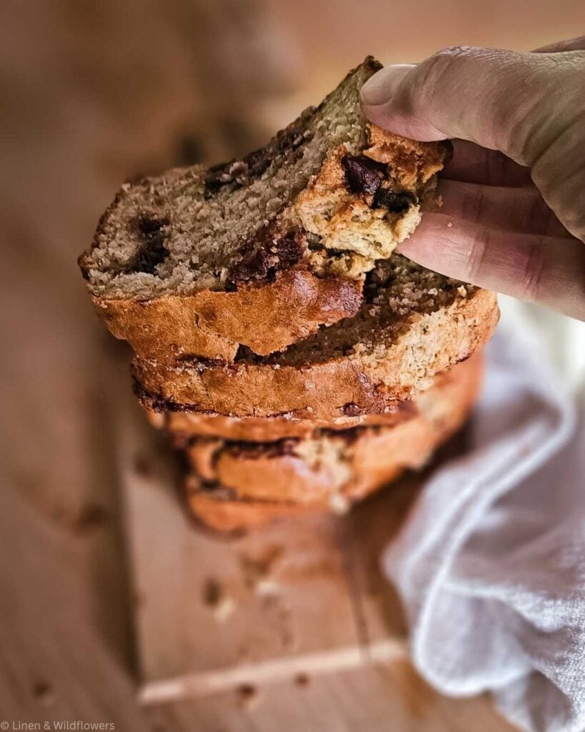 Close-up of a hand holding a slice of freshly baked sourdough banana bread with chocolate chips. The bread is moist and stacked on a wooden board, creating a warm, homemade feel.