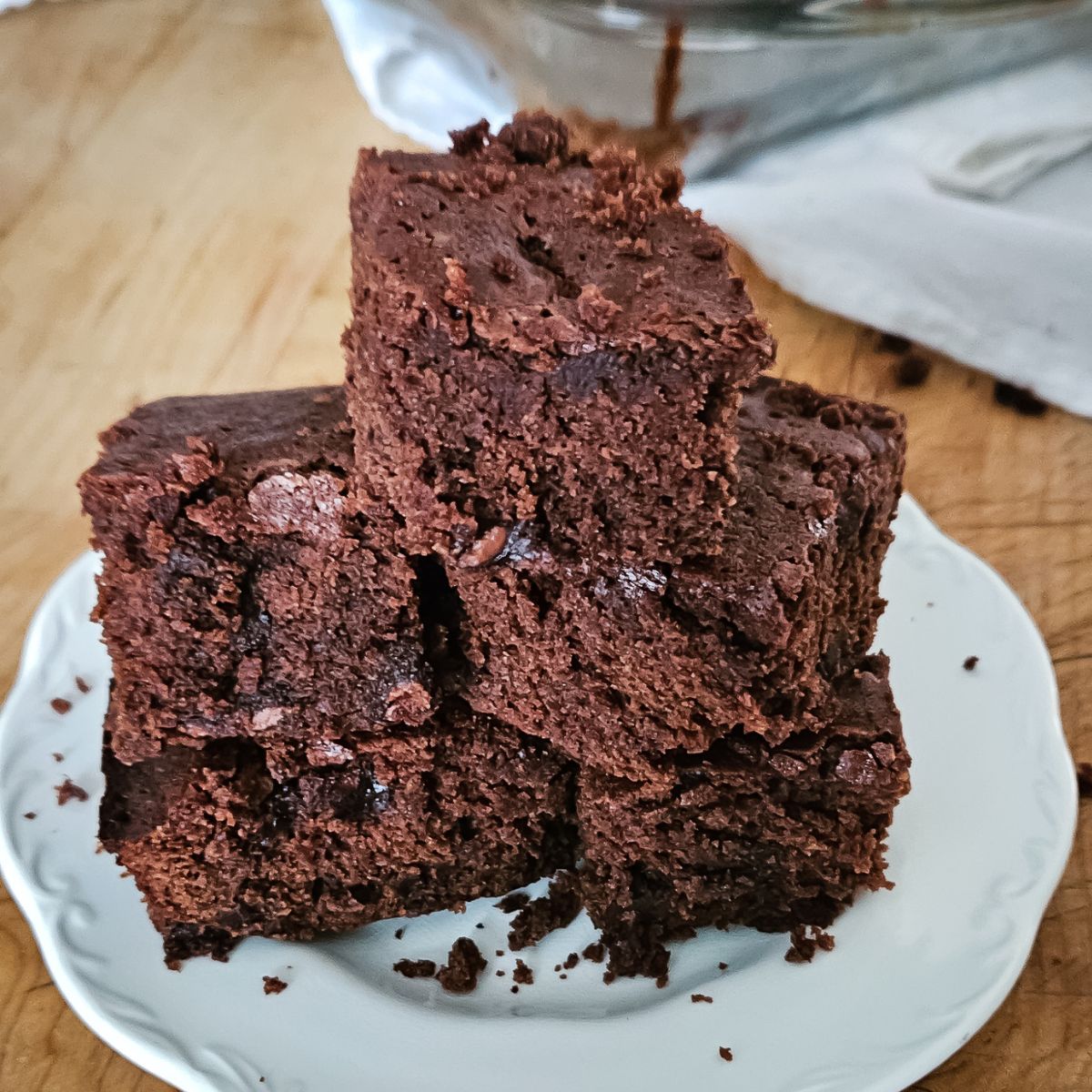 A stack of rich, chocolate brownies with a moist texture is arranged on a white plate, set on a wooden table. The scene feels warm and inviting.