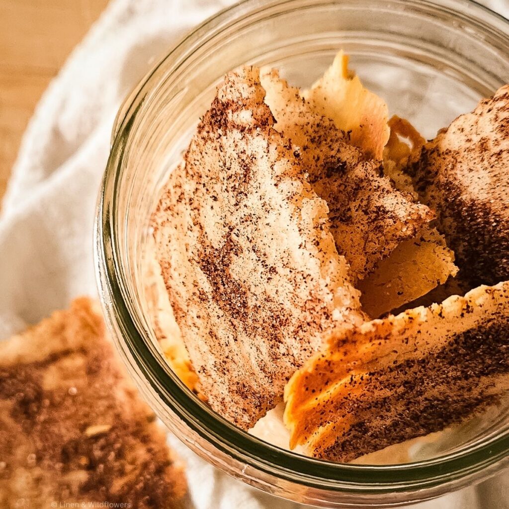 Close-up of crispy, brown sugar-topped crackers in a glass jar on a white cloth background, casting a warm and inviting feel.