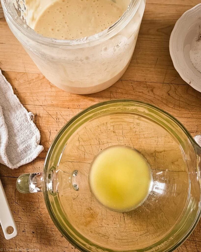 A wooden table with baking ingredients: a jar of creamy batter, a glass bowl with melted butter, a white bowl with flour, and a white cloth.