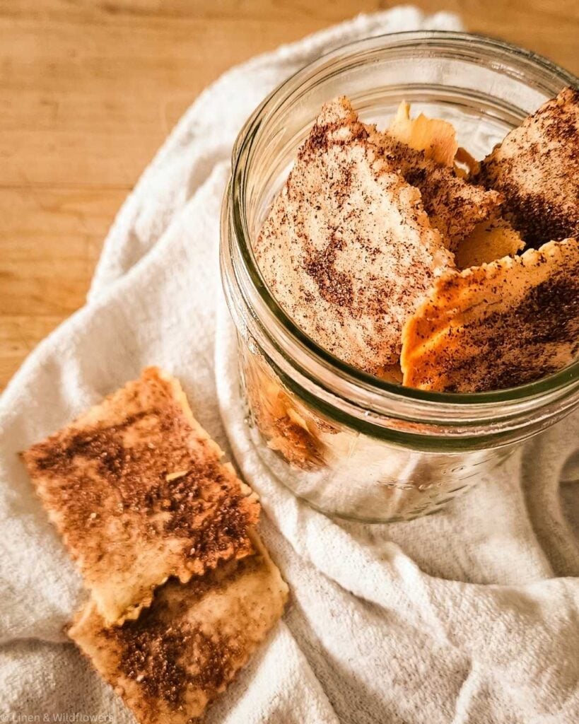 A glass jar filled with crispy, sourdough cinnamon-dusted crackers on a white napkin. Two additional crackers lie beside it on a wooden surface. Warm, rustic tone.