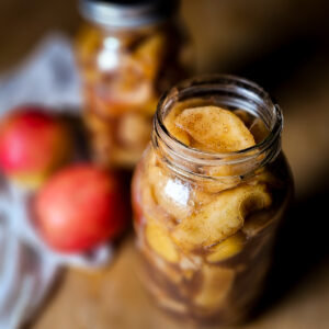A close-up of a glass jar filled with spiced apple slices, with a blurred background of another jar and two red apples. The image conveys a warm, homemade feel.