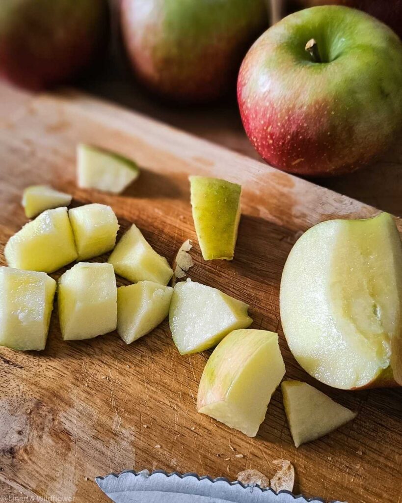 Sliced green apple pieces on a wooden cutting board beside whole apples, with a serrated knife in the foreground. Warm, fresh, and inviting.