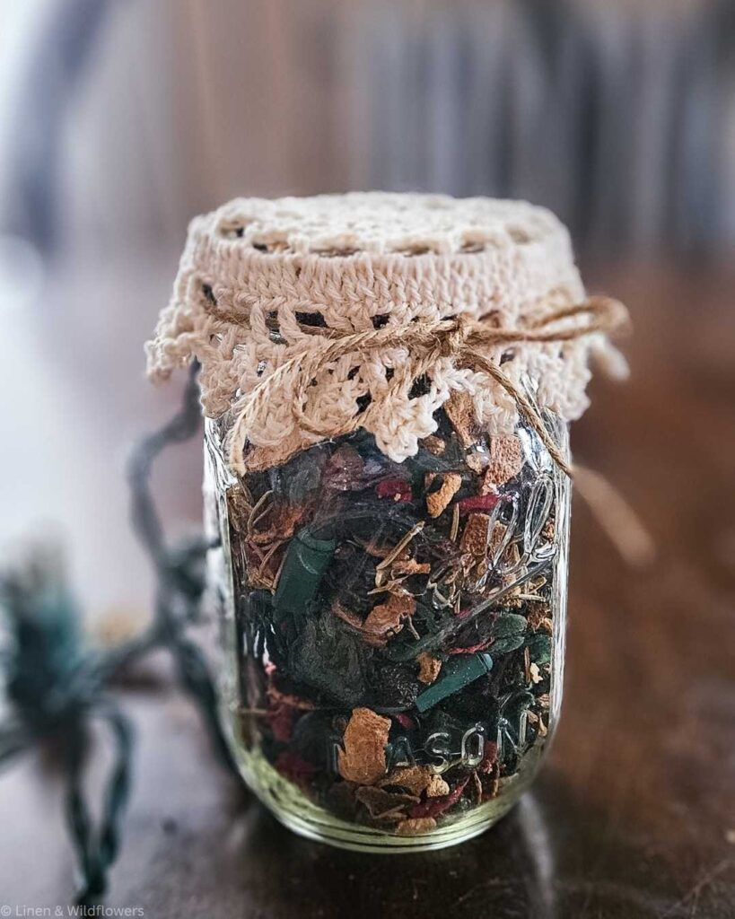 A Mason jar filled with potpourri, topped with a crochet lace cover tied with twine, sits on a wooden table. It conveys a rustic, cozy atmosphere.
