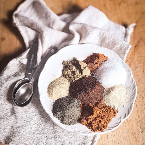 A white plate on a wooden table holds colorful spices, including brown sugar and coarse salt, arranged on a fabric napkin beside measuring spoons.