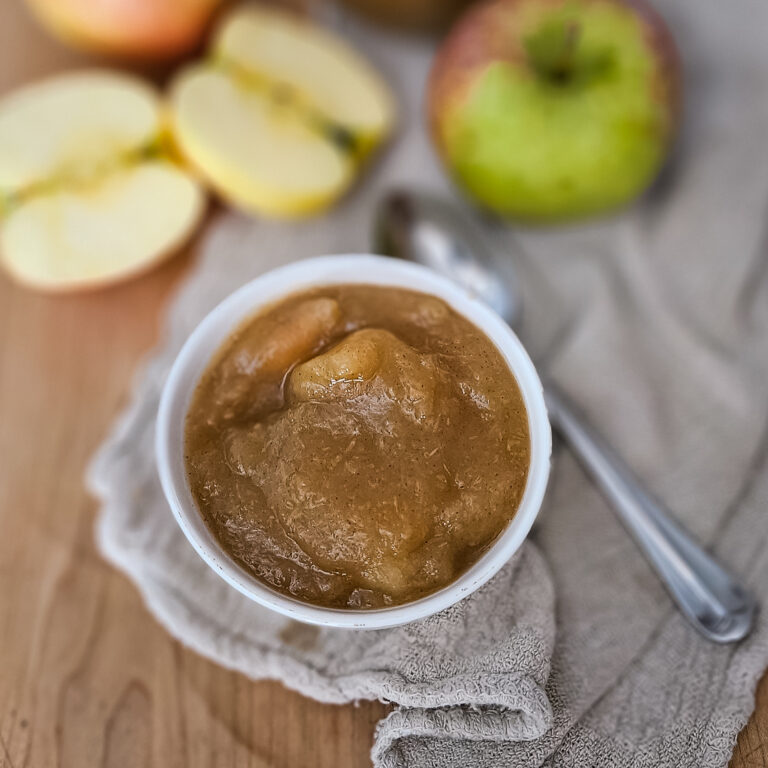 A bowl of chunky applesauce sits on a textured beige cloth beside a spoon. Fresh apples, both whole and sliced, rest on the wooden surface. The scene feels warm and inviting.