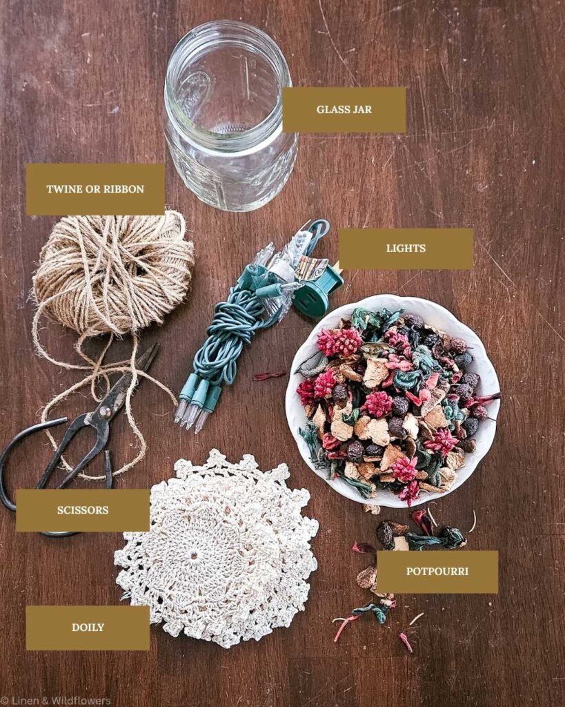 Craft supplies displayed on a wooden table: a glass jar, twine, scissors, string lights, a white doily, and a bowl of colorful potpourri. Cozy and creative.