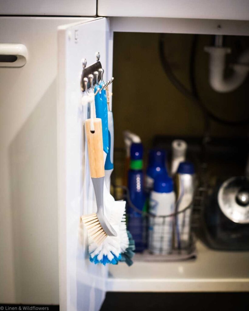 Under-sink cabinet open, showing hanging scrub brushes on hooks inside the door. Inside the cabinet are cleaning supplies in a basket. Bright and organized.