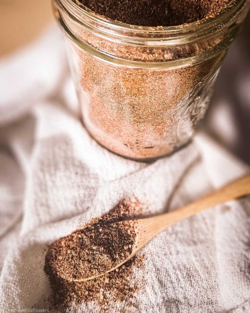 A glass jar filled with a chili seasoning spice mix sits on a white cloth. A wooden spoon lies across the cloth with some of the spice spilled beside it, evoking a rustic kitchen vibe.