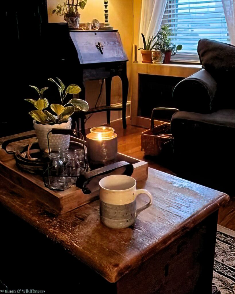 Cozy living room with warm lighting, featuring a lit candle and potted plant on a wooden table. A mug sits nearby. A vintage desk and window add charm.