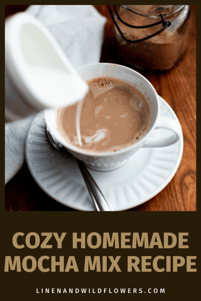 A cup of mocha with milk being poured in, set on a saucer with a spoon. A jar of cocoa powder is nearby. Text reads "Cozy Homemade Mocha Mix Recipe."