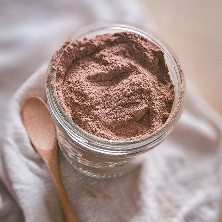 A glass jar filled with fine fireside mocha mix powder sits on a soft, folded beige cloth. A wooden spoon rests beside it, conveying a warm, cozy atmosphere.