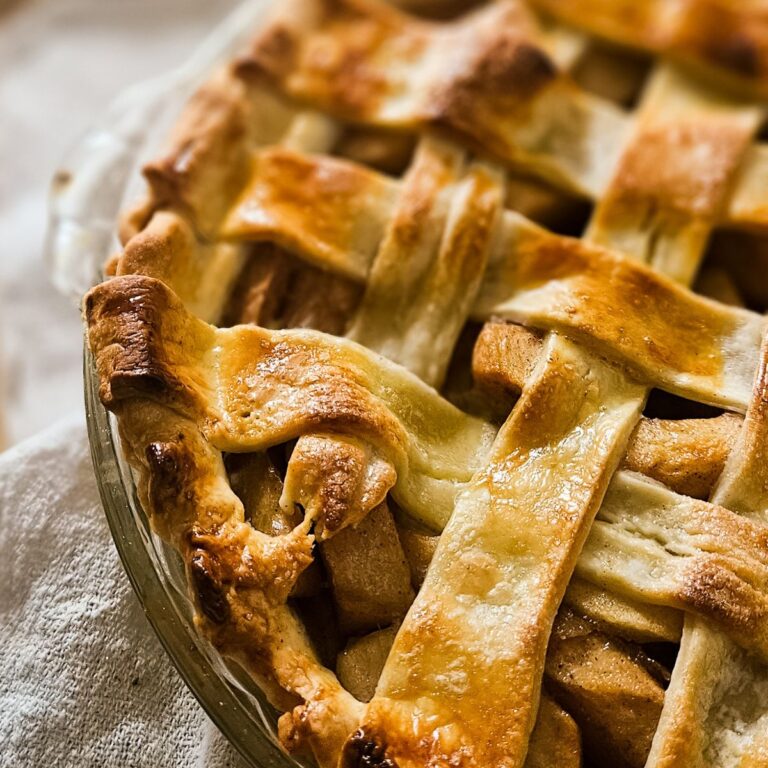 Close-up of a golden-brown, lattice-topped apple pie in a clear pie dish. The crust is flaky and shiny, conveying warmth and a homemade feel.
