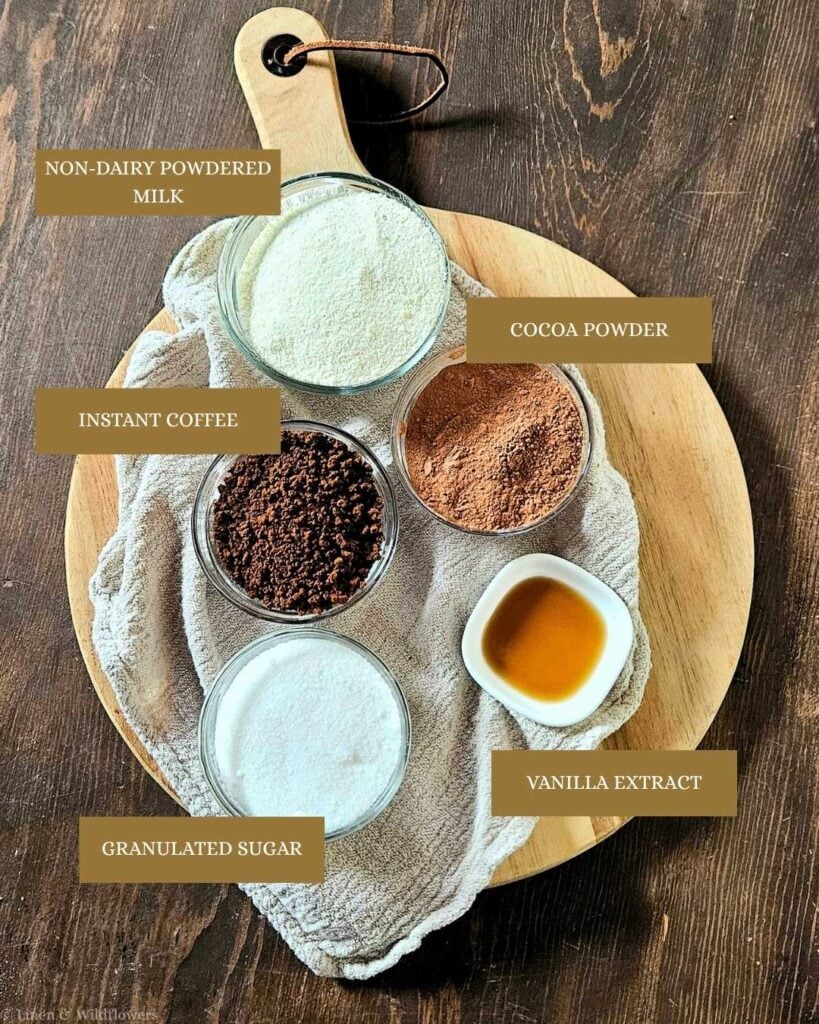A wooden board displays labeled ingredients: non-dairy powdered milk, cocoa powder, instant coffee, vanilla extract, and granulated sugar in bowls.