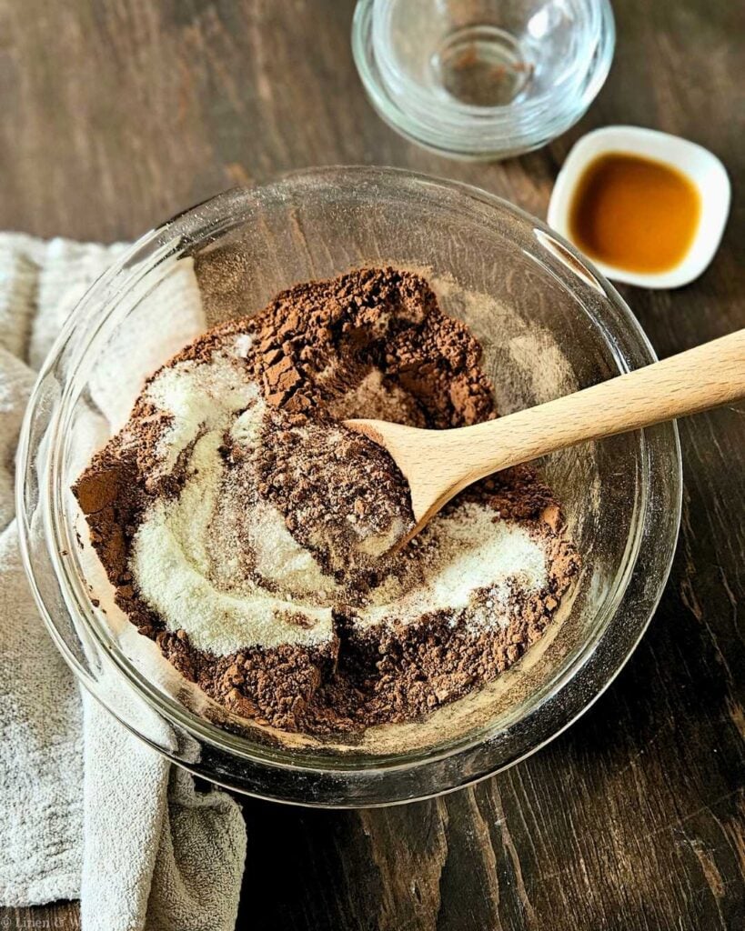 A glass bowl on a wooden table holds cocoa powder and flour, mixed with a wooden spoon. Nearby are a glass dish and a small bowl of vanilla. Baking setup.