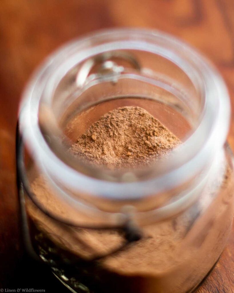 A glass jar, viewed from above, holds a pile of light brown cinnamon powder. The wooden background adds warmth, evoking a cozy, rustic feel.
