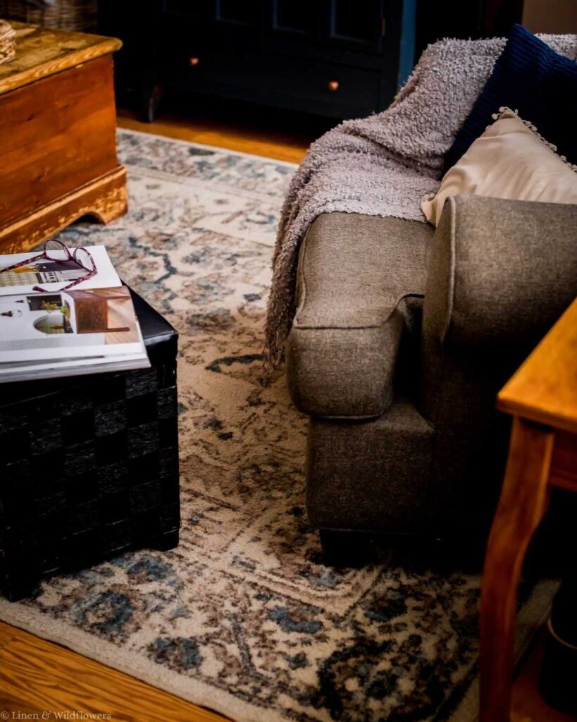 Cozy living room corner with a gray armchair draped in a soft blanket. A patterned rug covers the floor, and a wooden table holds magazines. Warm, inviting ambiance.